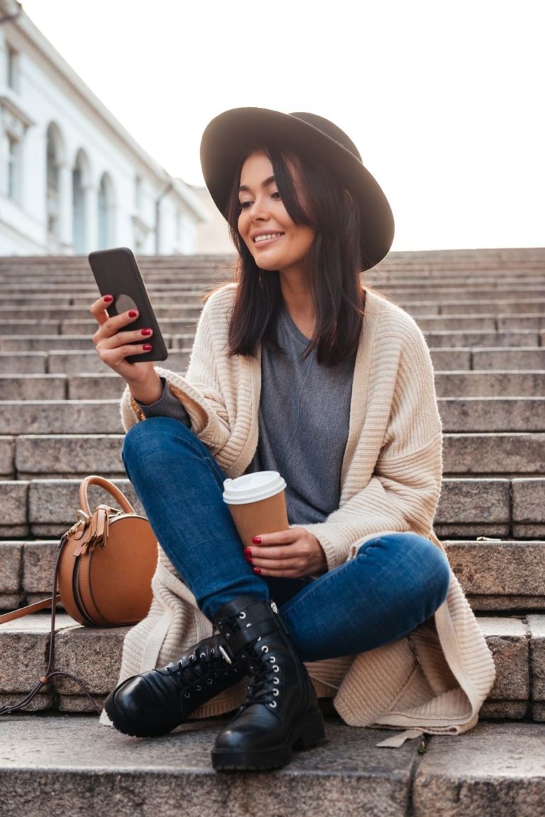 Girl is sitting on steps outside with coffee wearing a hat and cardigan, smiling, looking at her phone