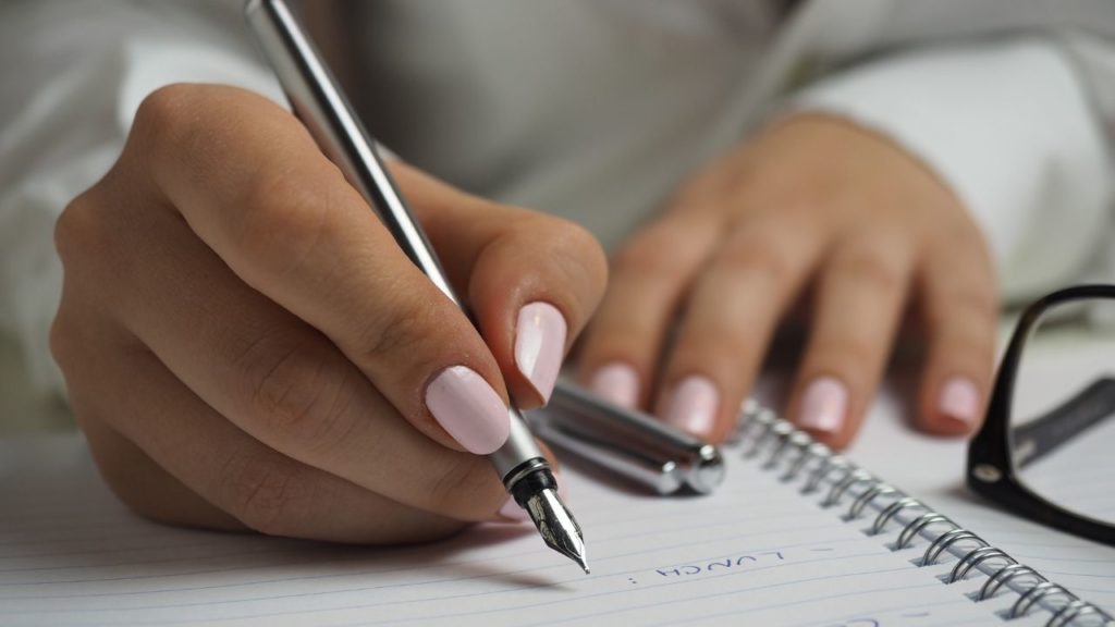 Woman with pretty pink fingernails is holding a pencil and is writing in a notbebook with a pair of glasses nearby