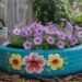 A vibrant blue upcycled tire planter, decorated with cheerful painted flowers and filled with blooming purple petunias, adds creative DIY flair to the garden. Surrounded by mulch, lush greenery, and a metal watering can, this eco-friendly project beautifully combines sustainability with eye-catching design.