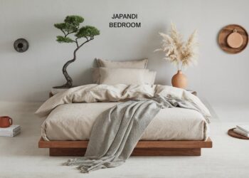 Minimalist Japandi bedroom with a low wooden bed, beige bedding, and a textured gray throw. A small tree and pampas grass decorates each side. A round wall shelf holds a wicker tray. Neutral tones create a serene atmosphere.