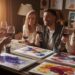 In a cozy room adorned with vibrant abstract paintings, four friends gather around a wooden table, wine glasses in hand and smiles on their faces. The warm lighting adds to the atmosphere, creating an inviting space perfect for sharing creative ideas and enjoying each others company.