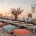 A bohemian-style outdoor dining setup at sunset exudes the ultimate Boho vibe. A low wooden table with colorful cushions, floral centerpieces, candles, and greenery graces the rooftop. String lights and a macramé wall hanging enhance this enchanting ambiance.