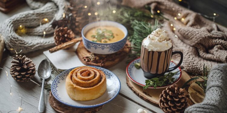 A cozy winter scene features a cinnamon roll on a plate, a mug of hot chocolate with whipped cream, and soul-warming soup. Surrounded by pinecones, knit scarves, and twinkling lights on a wooden table, its the perfect spread of cozy winter recipes to warm your soul.