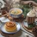 A cozy winter scene features a cinnamon roll on a plate, a mug of hot chocolate with whipped cream, and soul-warming soup. Surrounded by pinecones, knit scarves, and twinkling lights on a wooden table, its the perfect spread of cozy winter recipes to warm your soul.
