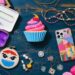 A flat lay of colorful crafting supplies on a blue wooden table, featuring yarn, beads, a cupcake toy, a smartphone with a colorful case, embroidery, containers, and thread. Bright and vibrant, showcasing a variety of creative tools.