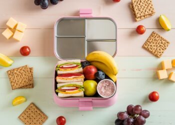 A pink lunchbox with sections containing sandwiches, a banana, an apple, blueberries, and yogurt. Surrounding the box are cubes of cheese, cherry tomatoes, apple slices, grapes, and crackers on a pastel background.