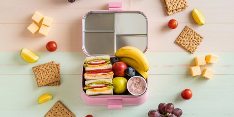 A pink lunchbox with sections containing sandwiches, a banana, an apple, blueberries, and yogurt. Surrounding the box are cubes of cheese, cherry tomatoes, apple slices, grapes, and crackers on a pastel background.
