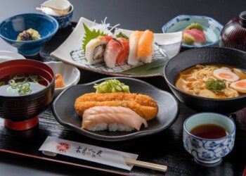 A Japanese meal set featuring sushi, noodle soup, pickled vegetables, miso soup, and tea on a black tray. The sushi includes nigiri and rolls, and a piece of fried tempura is placed over rice. The dishes are served in ceramic bowls and plates.