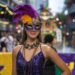 Woman in a purple sequin dress and feathered mask stands in front of a festive parade background. She holds a staff and wears layered necklaces. Colorful banners decorate the street, with blurred crowds and floats in the distance.