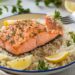 A plate of grilled salmon garnished with lemon herb sits on a bed of white rice. Lemon wedges are arranged around the edge. A fork with a bite of rice and salmon hovers above the plate, showcasing this delicious rice recipe. Parsley sprigs are in the background.