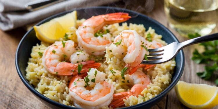 A bowl of garlic butter shrimp risotto garnished with parsley. Several shrimp rest atop the creamy rice. A fork is lifting one shrimp from the dish. Lemon wedges are on the side, and a glass of white wine is in the background, making this a perfect one-pan dinner delight.