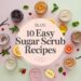 Text reading Blog: 10 Easy Sugar Scrub Recipes is surrounded by various colorful jars of sugar scrubs mixed with fresh fruits, herbs, and vanilla pods on a pastel pink and green background.