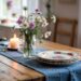 A sunlit, cozy room is warmed by farmhouse decor, featuring a wooden table adorned with floral plates, wildflowers, and a DIY table runner for inviting rustic charm.