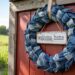 A charming DIY wreath made from repurposed denim and adorned with a “welcome home” sign adds a cozy, rustic touch to a red barn wall, set against the serene beauty of green fields and open skies.