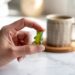 A hand lifts a vibrant Grüns Gummies candy beside a steaming mug on sunlit marble, embodying a simple moment of daily empowerment and joy.