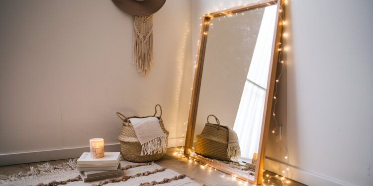 Transform your space with a cozy corner: a wood-framed full-length mirror adorned with string lights, complemented by woven baskets, candles atop stacked books, and stylish wall decor—making for an inspiring glow-up mirror setup.