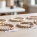 Discover six minimalist beaded bracelets in neutral tones—each with unique names like NEUTRAL VIBES and OCEAN WOOD—perfect for lovers of simple, stylish accessories. Displayed on a light wooden table against a bright, airy backdrop, these bracelets are must-haves for anyone embracing understated elegance.