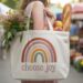 At a bustling farmers market, a woman with a DIY choose joy tote—adorned with a boho rainbow—shops for fresh flowers, peaches, and baguette amid vibrant stalls and colorful produce, capturing the spirit of positivity and mindful living.