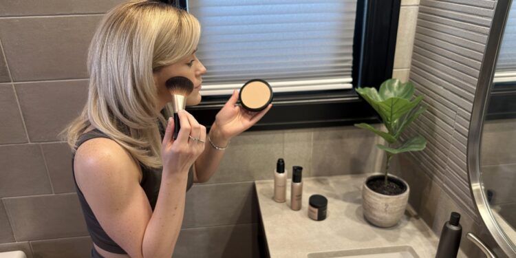 A woman perfects her beauty routine by applying Laura Geller Double Take Foundation in front of her bathroom mirror, surrounded by makeup essentials and a fresh potted plant—making every step feel enjoyable and worthwhile.