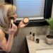 A woman perfects her beauty routine by applying Laura Geller Double Take Foundation in front of her bathroom mirror, surrounded by makeup essentials and a fresh potted plant—making every step feel enjoyable and worthwhile.