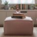 A pink ottoman with built-in storage serves as a stylish focal point in a cozy bedroom filled with plants, books, and personal decor, enhancing both function and ambiance beneath sunlit sheer curtains.