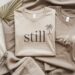 Two stylish, neutral-toned t-shirts—each featuring soothing palm tree designs and the words still and calm—are artfully arranged alongside chic accessories like gold earrings, a coffee cup, and a dried palm leaf, evoking a serene vibe perfect for effortless, mindful living.