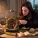 In her warmly lit study, a woman finds inspiration at a desk lined with books, captivated by a birdcage reimagined as an enchanting DIY Book Nook. Inside, miniature shelves and tiny figurines create a magical library scene—inviting creativity, imagination, and the wonder of literary worlds into her everyday life.