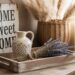 A charming thrifted vignette features a floral ceramic jug and fresh lavender, set atop a wooden tray. Rustic touches—like pampas grass in a wicker basket and a “Home Sweet Home” sign glowing in natural light—capture the cozy essence of curated secondhand decor.