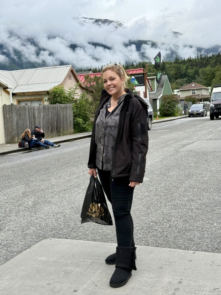 A woman, dressed in her DFIWE0886-list jacket and boots, smiles with a black shopping bag on a street lined by homes and misty hills.