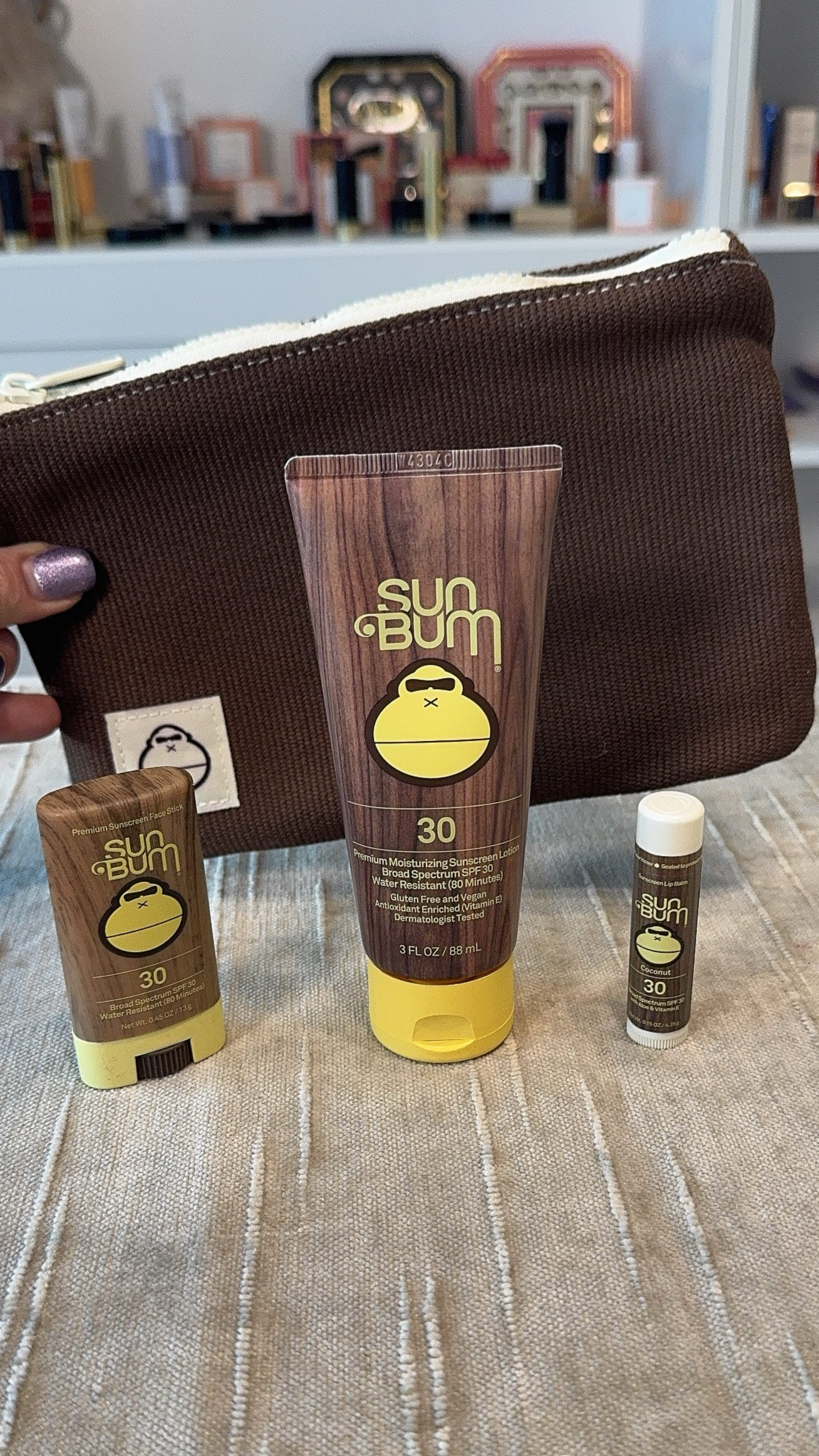Sun Bum toiletry bag and SPF 30 stick, tube, and lip balm rest on beige fabric—perfect for a cruise—skincare shelves behind.
