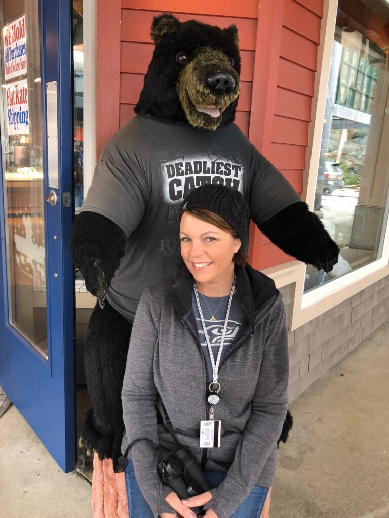 Grinning in a gray hoodie, a woman poses with IMG_1352—the bear statue—near a red building on an unforgettable Alaska trip.
