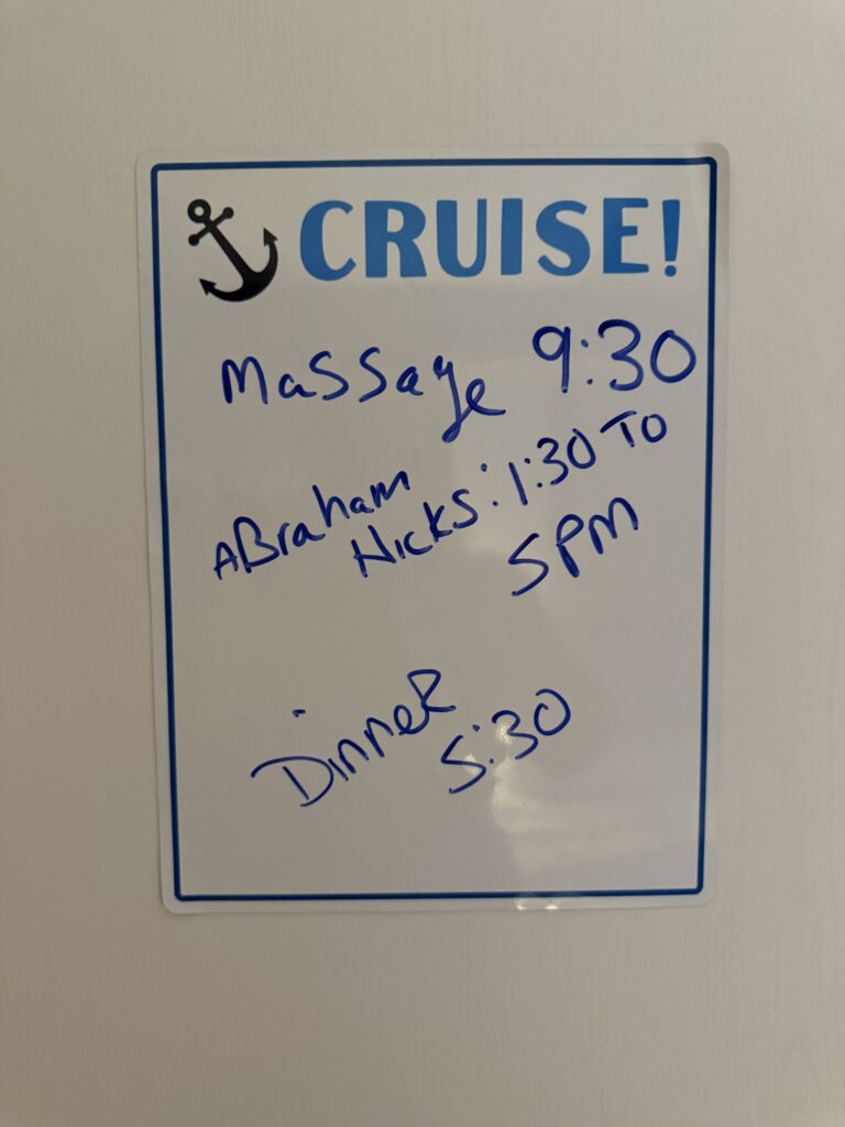 A whiteboard displays a blue anchor, “CRUISE!” and a handwritten schedule: Massage 9:30, Abraham Hicks 1:30–5, Dinner 5:30.