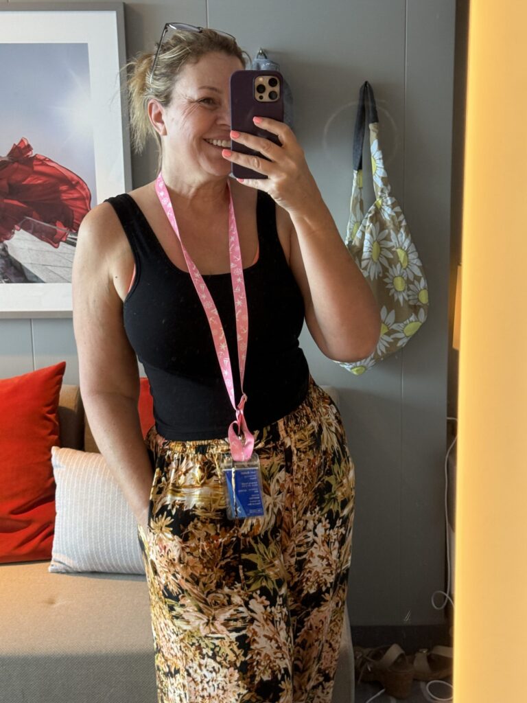 A woman beams in a mirror selfie, sporting a black tank, floral pants, and pink lanyard—her vibrant outfit perfect for a cruise.