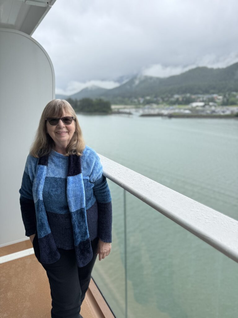 On a ship deck against misty, forested Alaskan mountains, a woman in chic blue attire and sunglasses embodies cruise style.