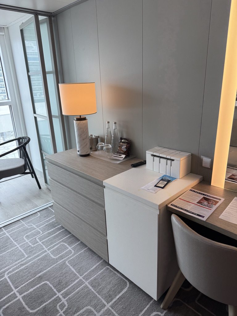 A sleek desk area with lamp, bottles, snacks, and paperwork sits by a windowed wall, perfect for cruise planning.