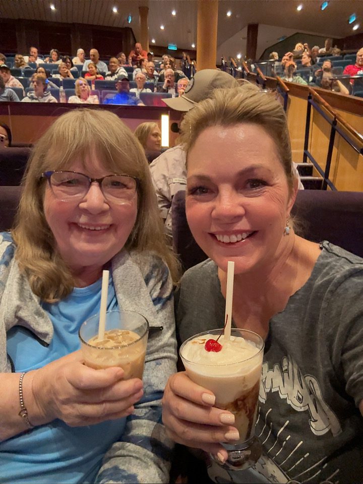 Smiling women sip cherry-topped cocktails in a vibrant cruise ship theater, showcasing onboard entertainment on B84F8EAD-2CE9.