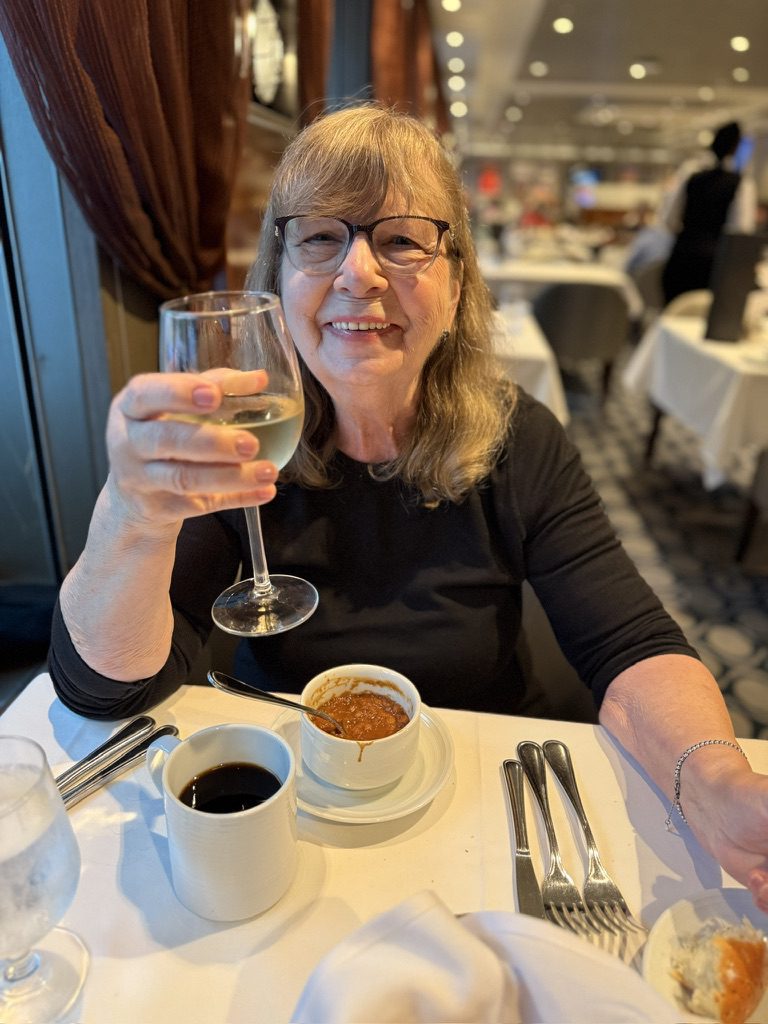 An older woman in glasses sips white wine at a bustling restaurant, radiating the laid-back vibe of cruise ship dining.