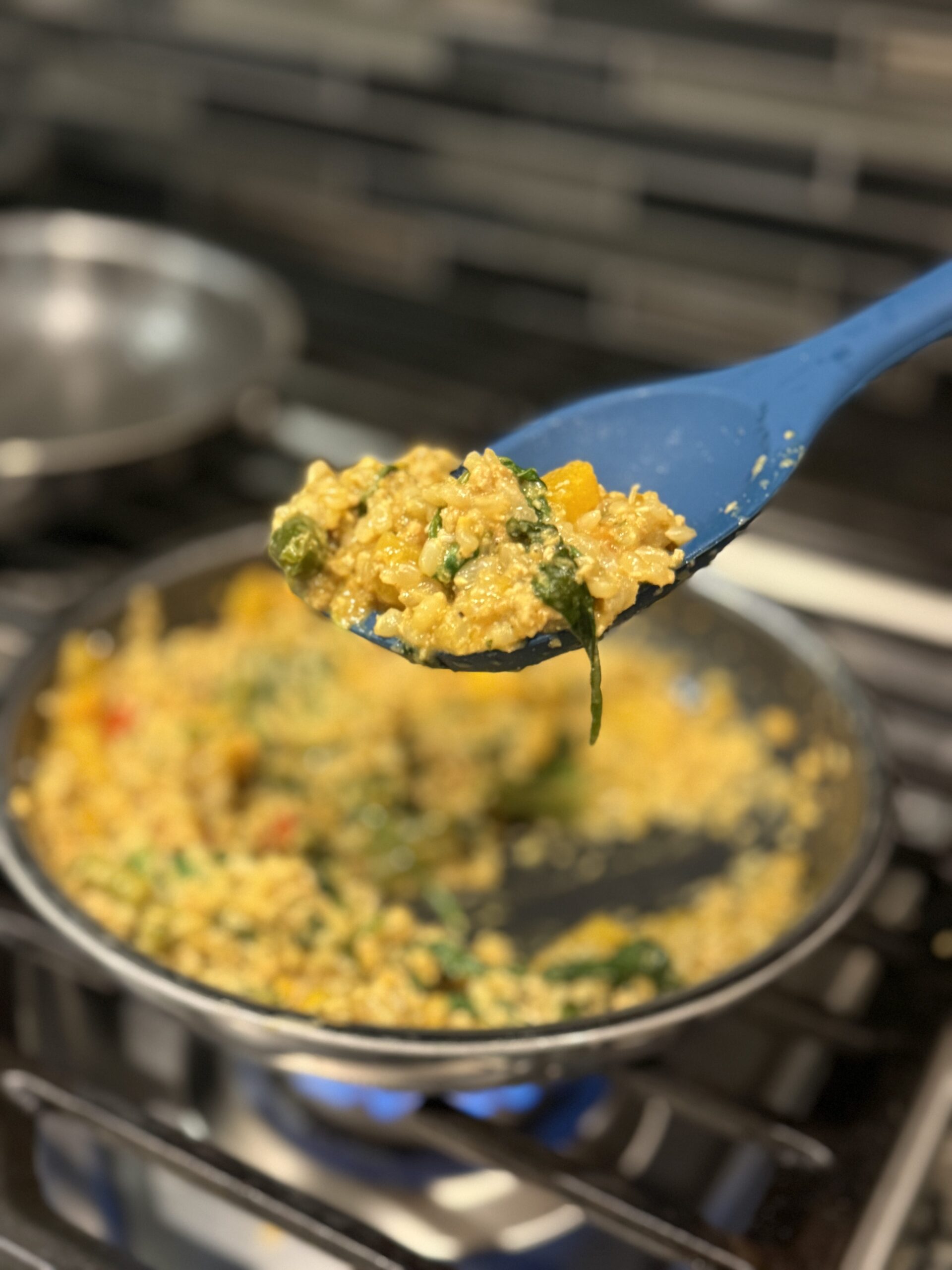 A blue spoon scoops vibrant vegetable rice from a pan on the stove, while another nonstick pan cooks in the background.