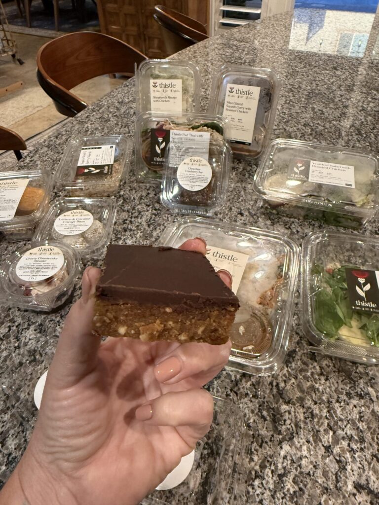 A hand displays IMG_1040, a chocolatey dessert bar, over a granite counter featuring Thistle meals and salads in the background.
