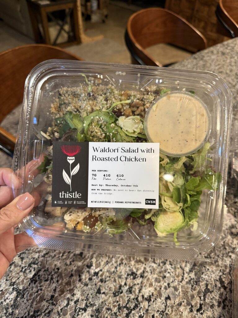 A hand grips a labeled plastic container (IMG_1194) filled with greens, nuts, chicken, and dressing—perfect protein for 50+.