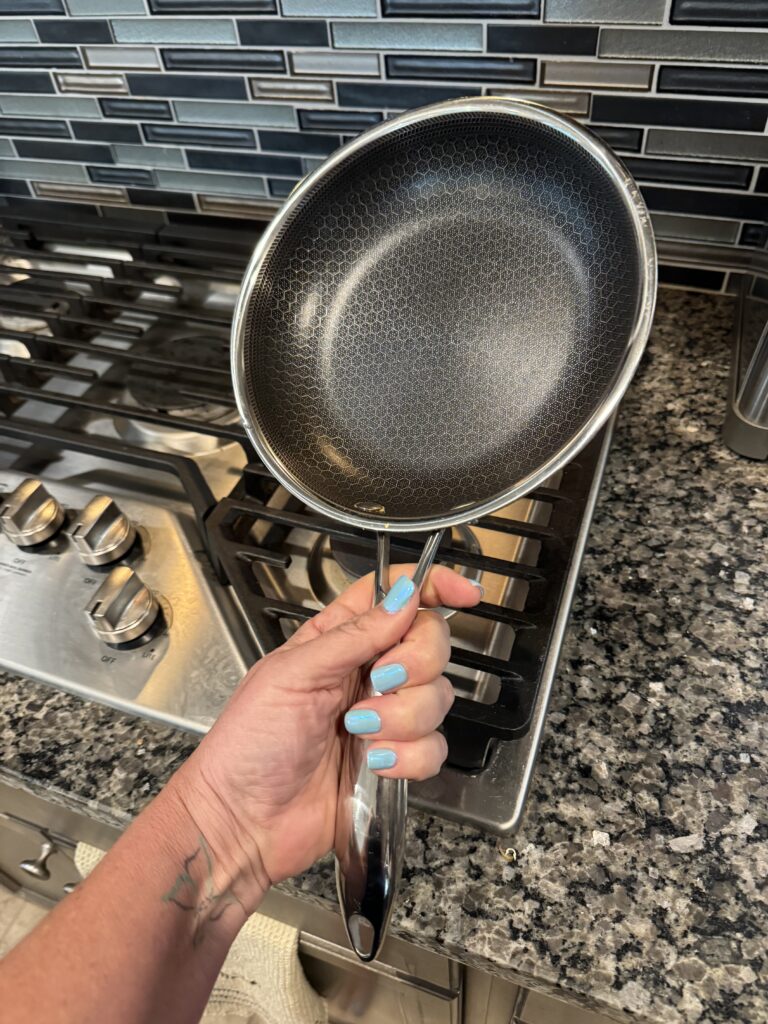 A person with pastel blue nails lifts IMG_2456—an easy-clean frying pan—over a stove set in a sleek, modern kitchen.