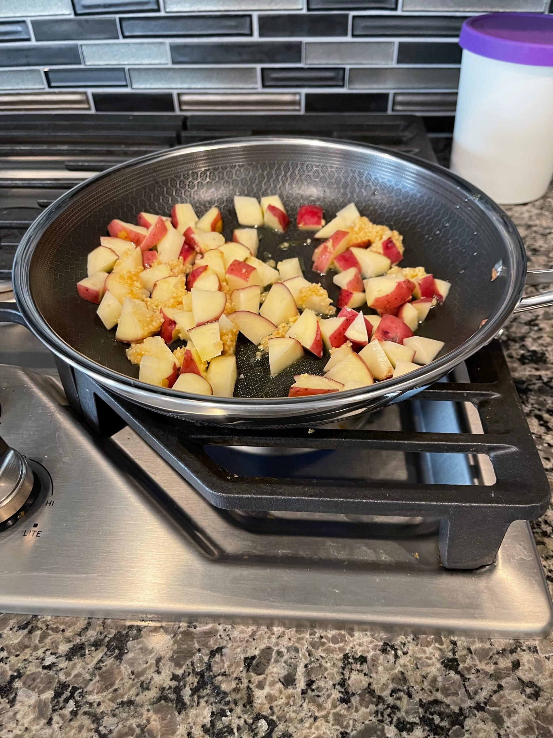 Sliced red potatoes and minced garlic cook in a nonstick skillet on a gas stove, against a tiled backsplash and container.