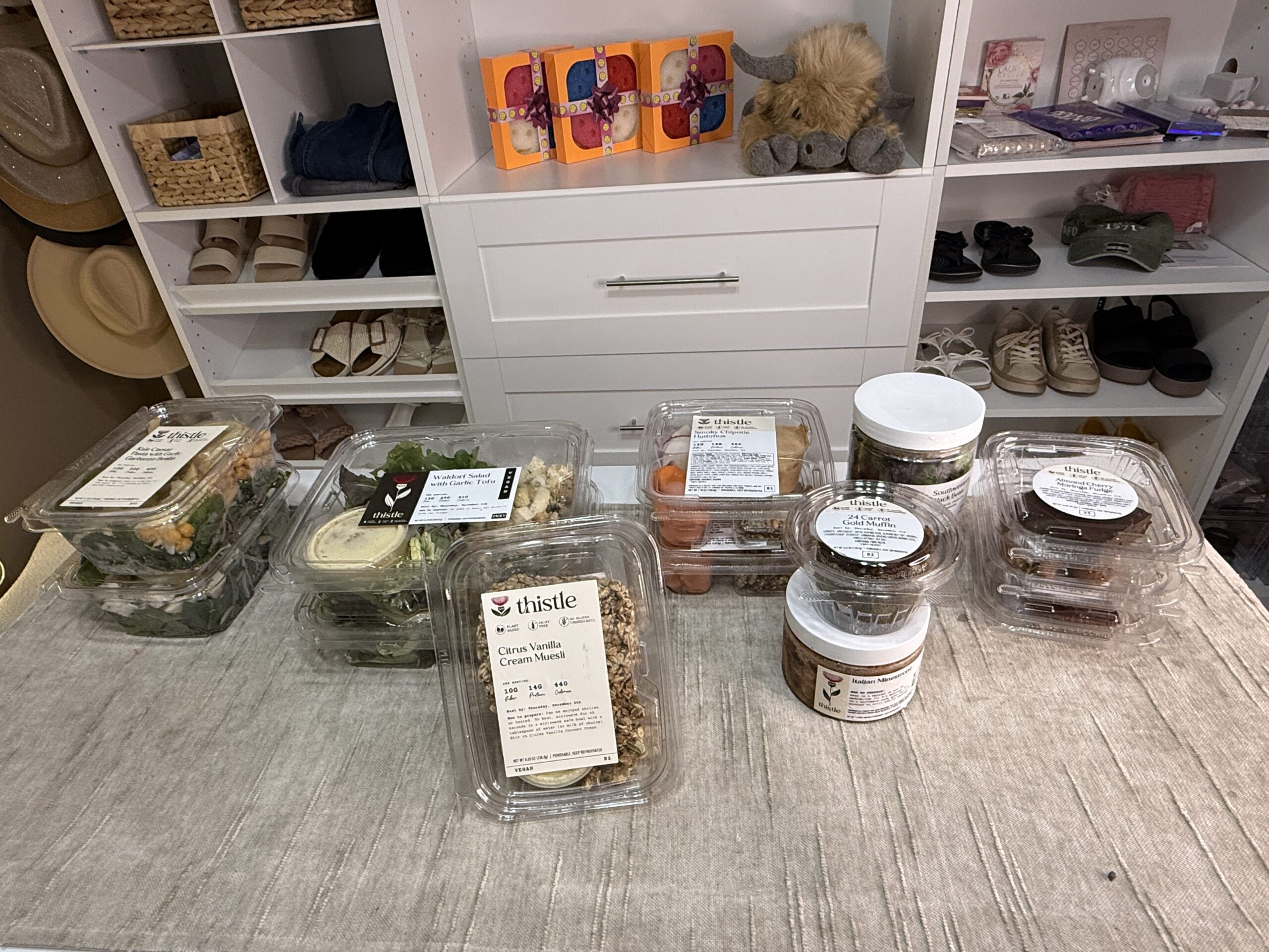 Assorted high-protein meals and snacks displayed on a beige cloth in a closet, with shelves stocked with shoes, hats, baskets, and blankets.